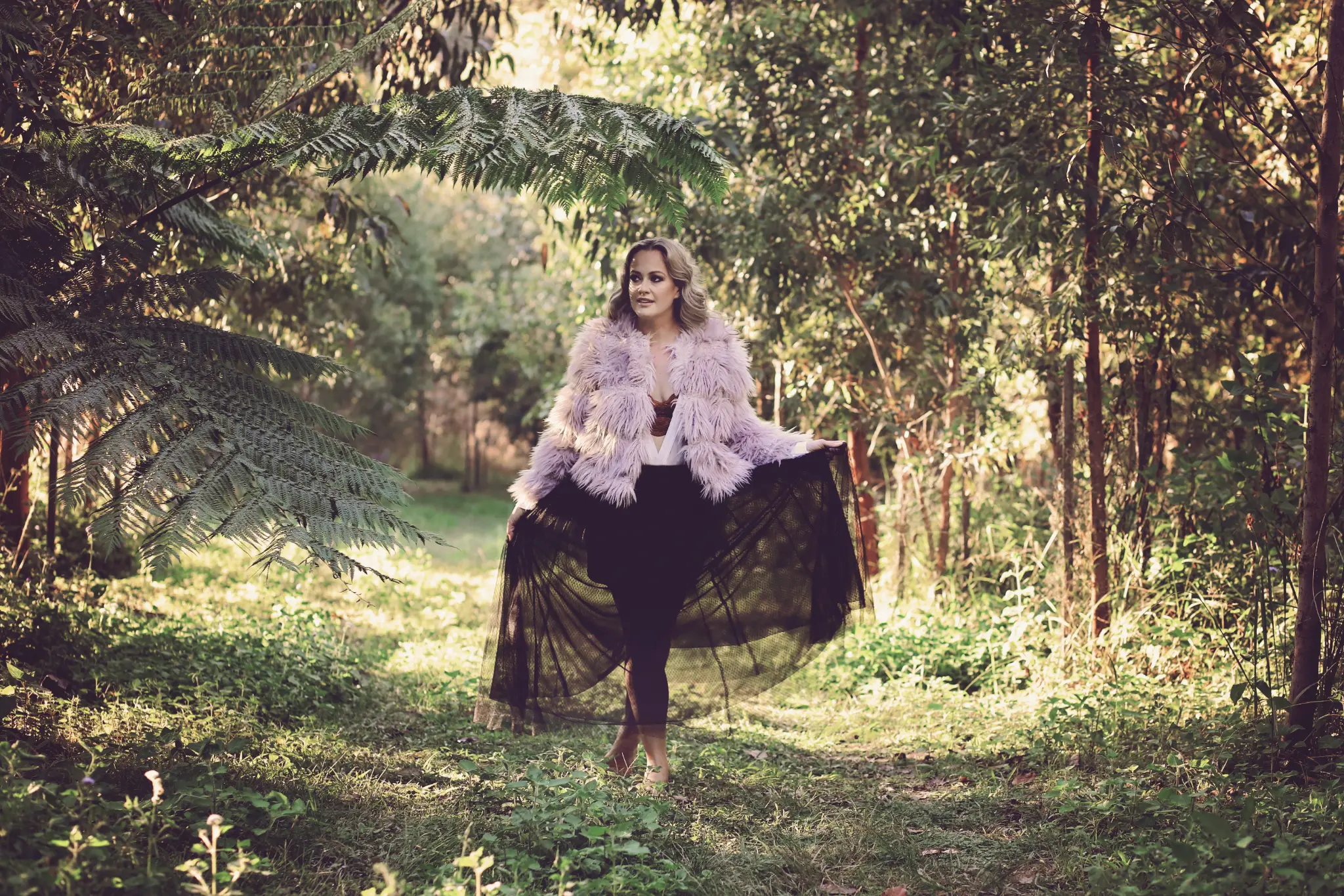 Lissy Katriona, in a flowing tulle skirt and lavender coat in a fern-lined bushland path